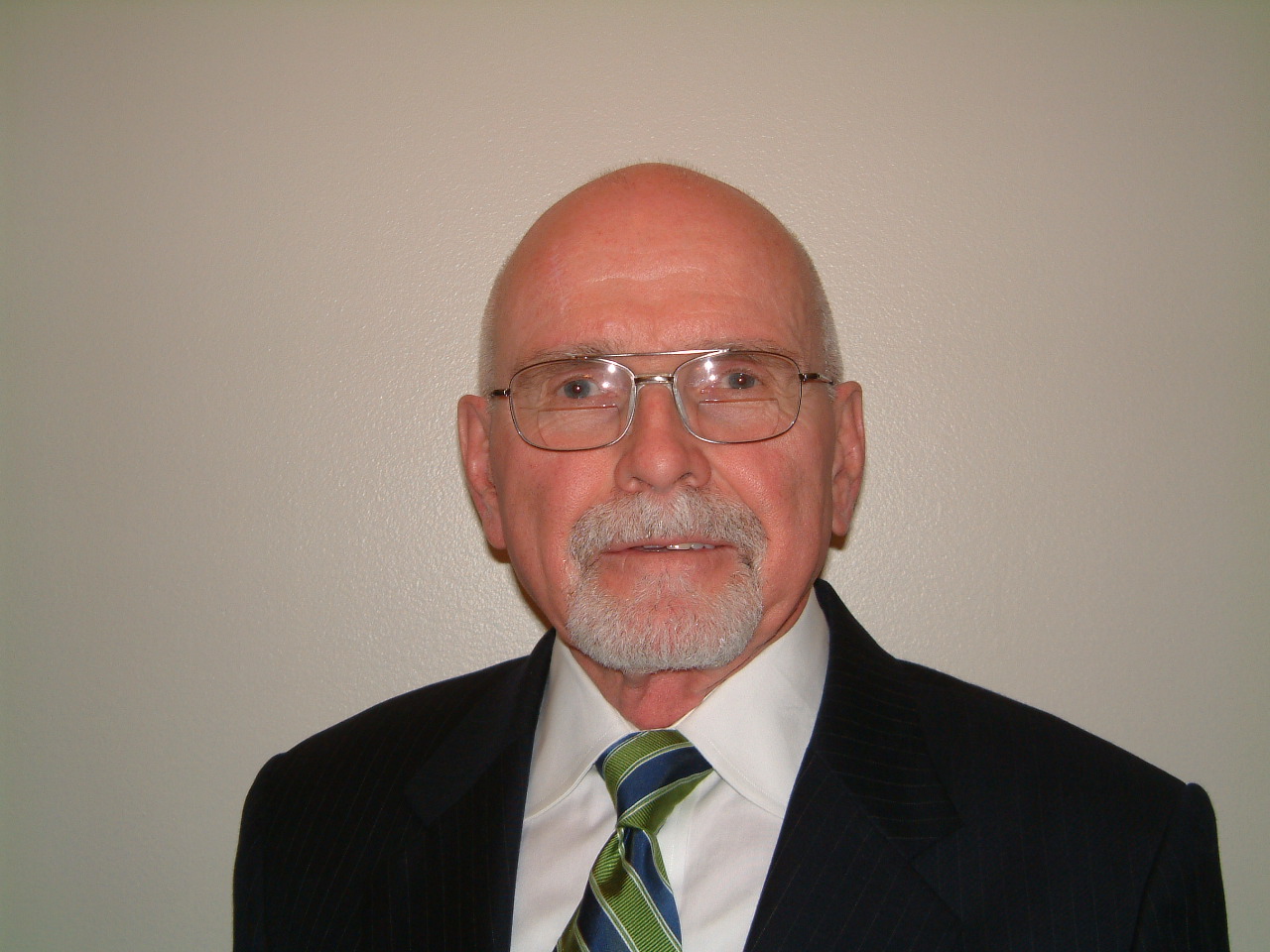 An older man in a suit and striped tie, wearing glasses, stands against a plain wall.