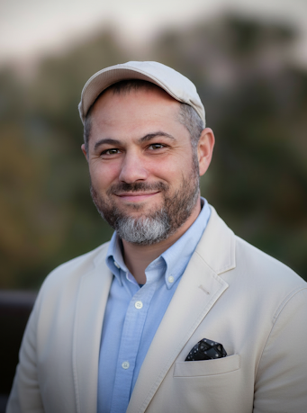 Headshot of a smiling man with a beard, wearing a flat cap, light blazer, and blue shirt.