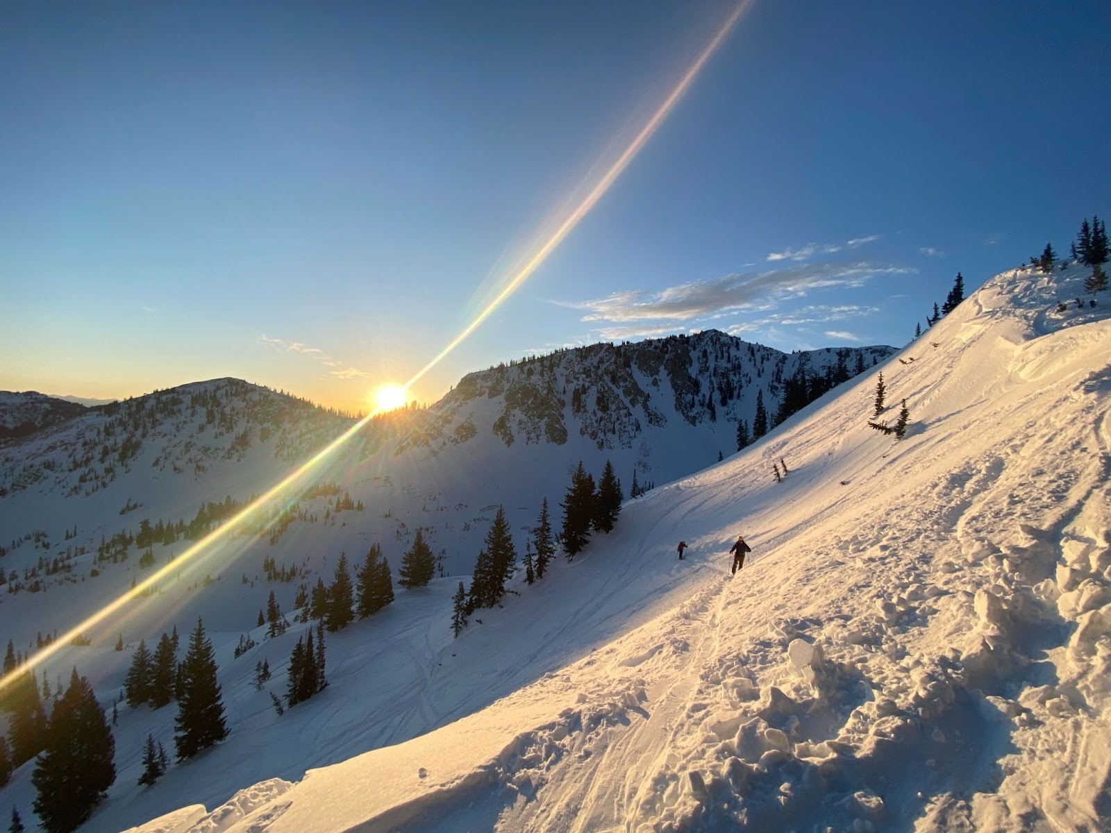 Sunset over snowy mountains with skiers descending a slope and trees scattered around